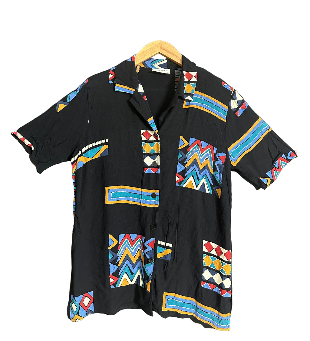 A black short-sleeve button-up shirt with colorful geometric tribal patterns, hanging on a wooden hanger against a dark gray wall.