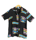 A black short-sleeve button-up shirt with colorful geometric tribal patterns, hanging on a wooden hanger against a dark gray wall.