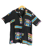 A black short-sleeve button-up shirt with colorful geometric tribal patterns, hanging on a wooden hanger against a dark gray wall.